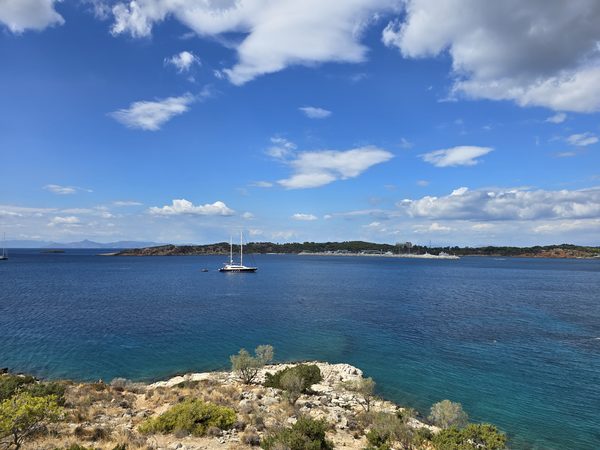 A rocky headland along the Athens Riviera coastline, looking out over a deep blue bay with a moored yacht