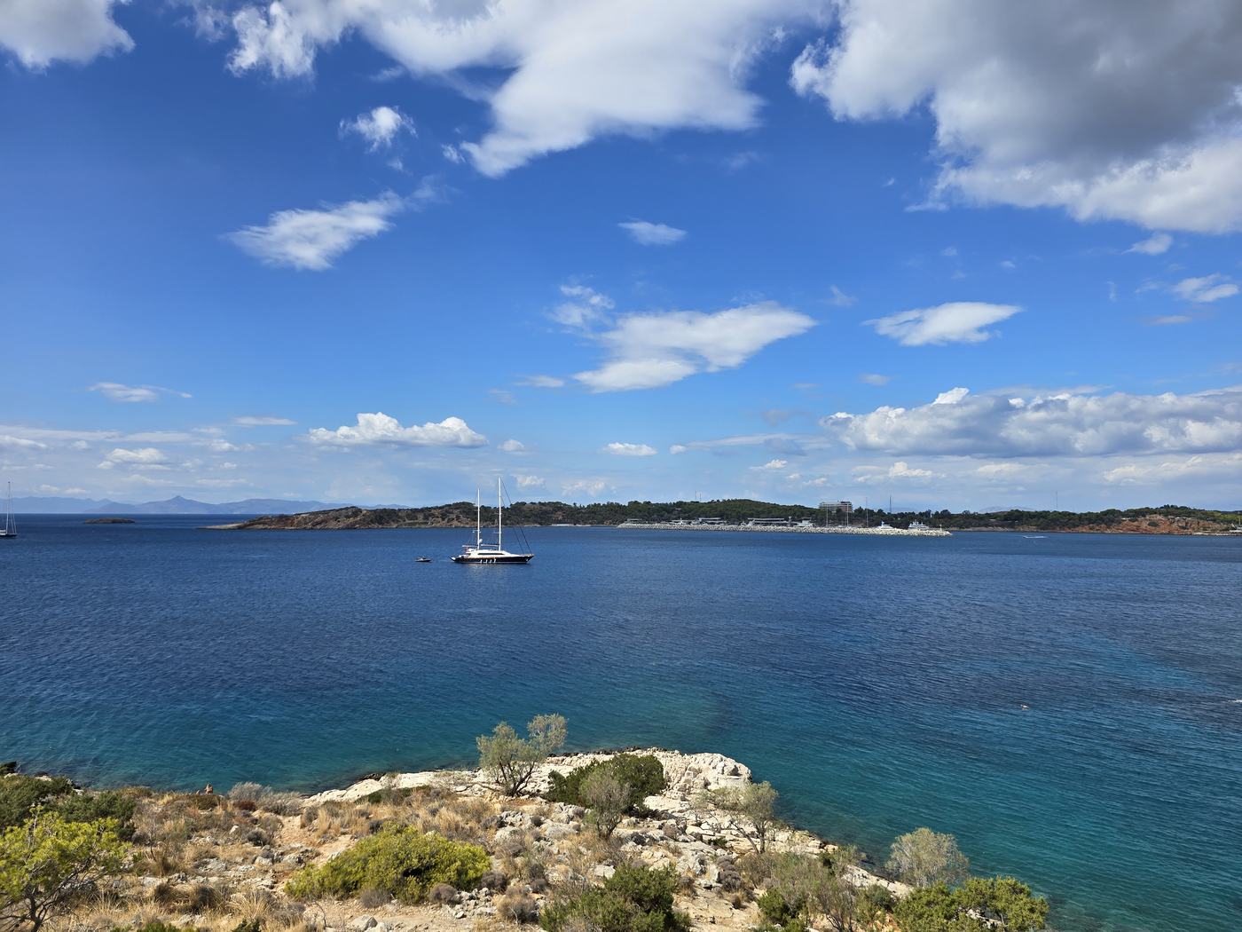 A rocky headland along the Athens Riviera coastline, looking out over a deep blue bay with a moored yacht