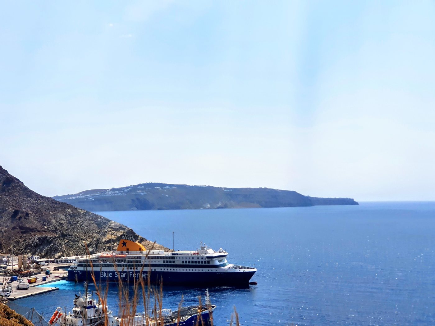 A Blue Star ferry docked at Santorini's Athinios port, with the switchback cliff road above and Thirasia island across the water