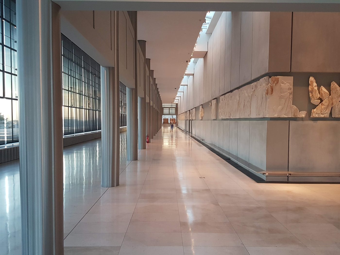 A long gallery inside the Acropolis Museum with Parthenon frieze marbles displayed along the wall and floor-to-ceiling glass looking out over Athens