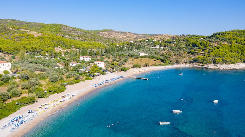 Aerial photograph of Agioi Anargyroi Beach on Spetses island, with turquoise water, a long pebbly shoreline, and pine-covered hills behind
