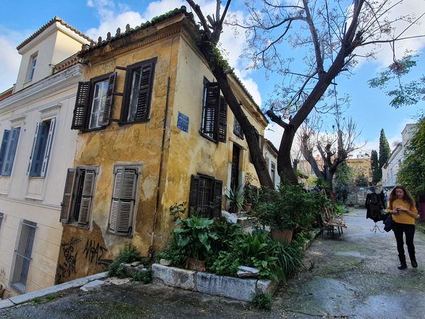 A quiet, ordinary street in the Plaka neighborhood with a pastel-colored old house, shuttered windows, and a pedestrian walking past
