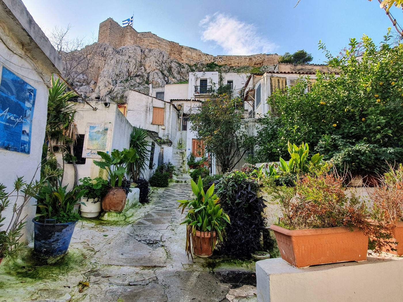 A narrow stepped lane in the Anafiotika neighborhood, lined with whitewashed houses and potted plants, climbing toward the Acropolis rock